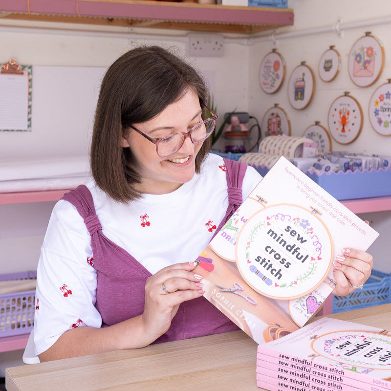 Woman holding a book titled 'sew mindful cross stitch' in a craft room with shelves and patterns in the background.